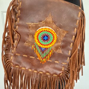 May include: A brown leather crossbody bag with fringe detailing. The bag features a beaded design with a circular pattern in shades of red, yellow, green, blue, and white.