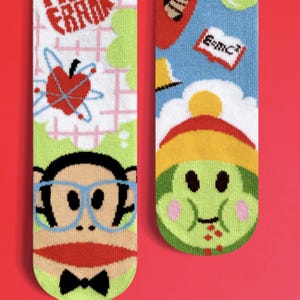 May include: A pair of colourful socks with a cartoon monkey wearing glasses on one sock and a green creature with a red mouth on the other sock. The socks have a school theme with images of an apple, a book, and a pencil.