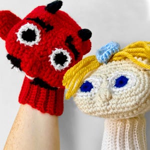 May include: Two handmade crochet puppets. One is red with black accents and white eye details. The other is off-white with yellow hair and blue eyes. A blue bow adorns the puppet's hair. The puppets are designed to fit over a hand.