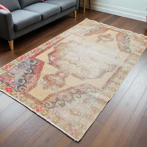 Puede incluir: Una alfombra rectangular de estilo vintage con un campo central beige y bordes ornamentados. La alfombra presenta un diseño desgastado con patrones descoloridos en rojo, azul y floral. Se coloca sobre un suelo de madera oscura, sugiriendo un salón.