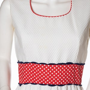 May include: A white sleeveless dress with a red and white polka dot band around the waist. The dress has a scalloped edge and a white background with a textured pattern.