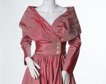 Può includere: Un abito lungo a maniche lunghe di colore rosa con scollo drappeggiato e gonna ampia. L'abito ha un corpetto aderente con una cintura e bottoni dorati.