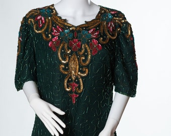 Può includere: Un abito di paillettes verde scuro con un motivo floreale colorato intorno al collo. L'abito ha maniche corte e una silhouette dritta.