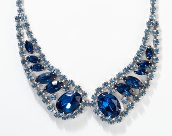 Può includere: Una collana vintage con una catena in metallo color argento ornata di strass blu chiaro e blu scuro. La collana ha un design a pettorina con grandi pietre ovali e a forma di marquise, creando un look scintillante ed elegante.