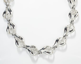 Può includere: Una collana argentata con un motivo a forma di fiamma ripetuto. La collana presenta una serie di forme a fiamma stilizzate e interconnesse, creando un motivo continuo. La catena è regolabile e ha una chiusura a moschettone.