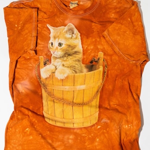 May include: Orange tie-dye t-shirt featuring a graphic of a ginger kitten sitting in a wooden bucket. The t-shirt has the text "The Mountain" printed on the bottom right corner.