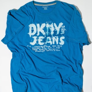 May include: A blue t-shirt with a white graphic print that says "DKNY JEANS" and includes the text "Lower East Side Est. 1989 New York City 24 hr. a day Soho Tribeca Chelsea Brooklyn China Town".