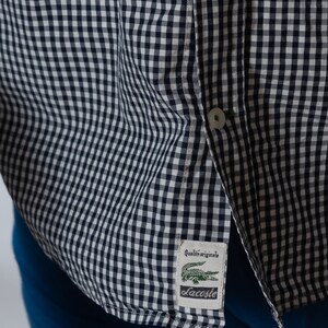 May include: Close-up of a navy and white checkered button-down shirt. The shirt features a small label with a green crocodile logo and the text "Qualit&eacute; originale" and "Lacoste". The shirt is partially unbuttoned.