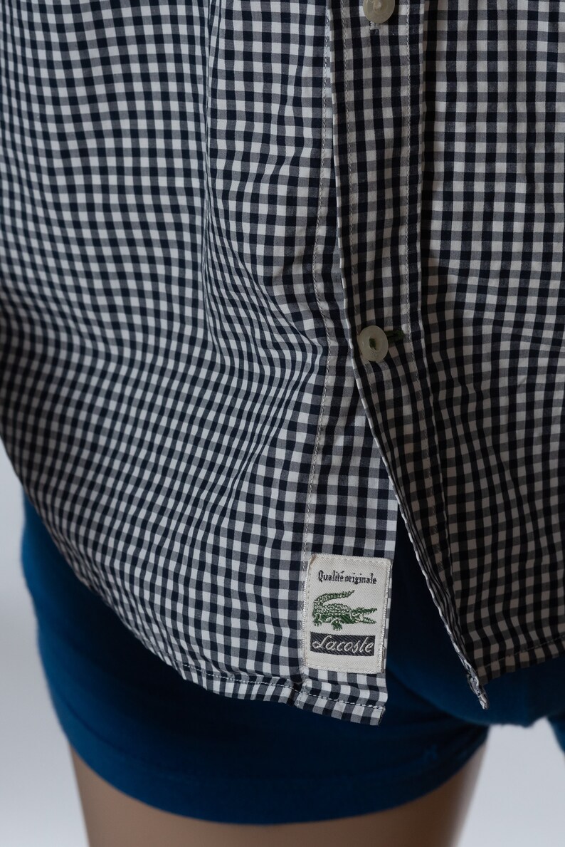 May include: Close-up of a navy and white checkered button-down shirt. The shirt features a small label with a green crocodile logo and the text "Qualit&eacute; originale" and "Lacoste". The shirt is partially unbuttoned.