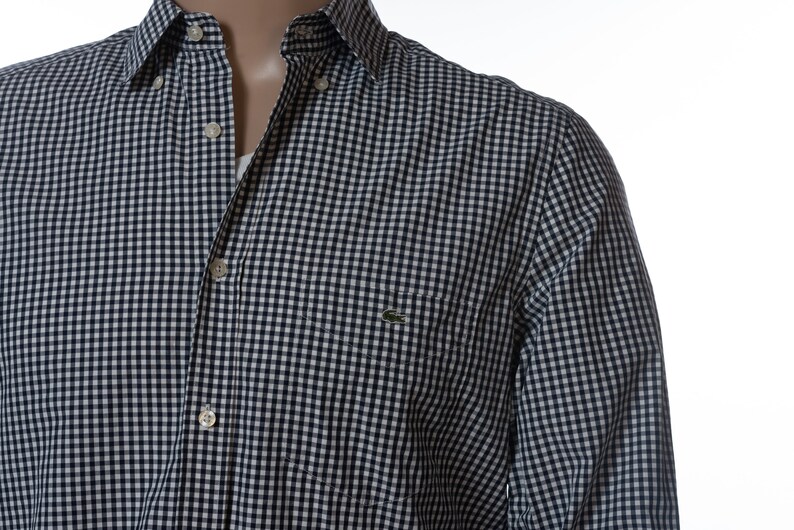 May include: A button-down shirt with a navy and white checkered pattern. The shirt has a small pocket on the chest and a green crocodile logo. The collar is buttoned down, and the shirt is partially unbuttoned.