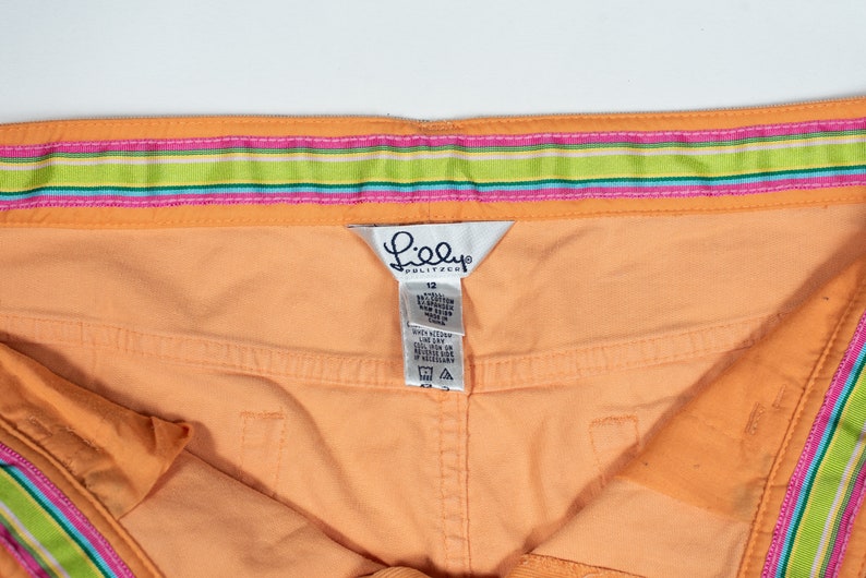 May include: Orange shorts with a pink, green, and blue striped waistband. The shorts have a Lilly Pulitzer tag with the size 12 printed on it.
