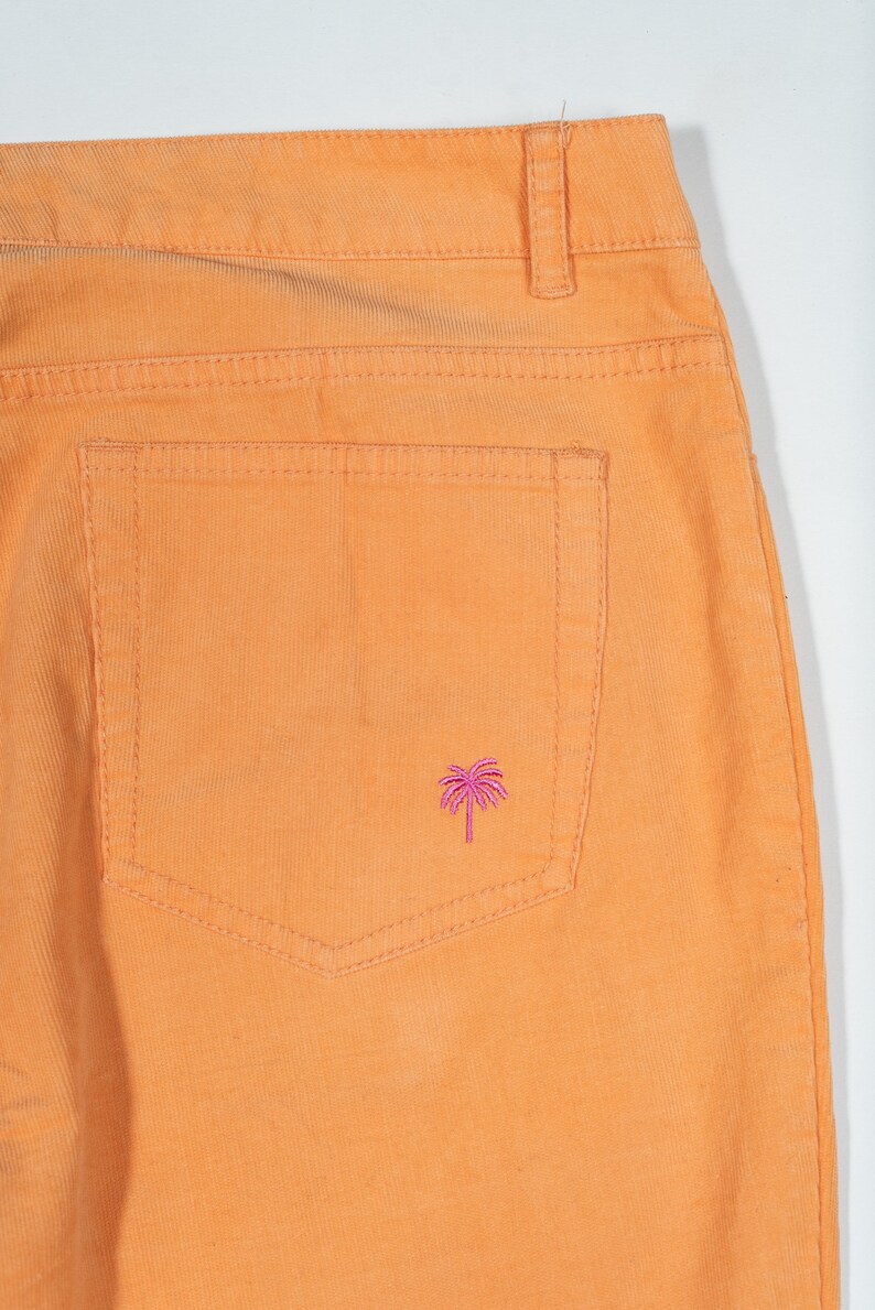 May include: Orange corduroy pants with a back pocket featuring a pink palm tree embroidery.