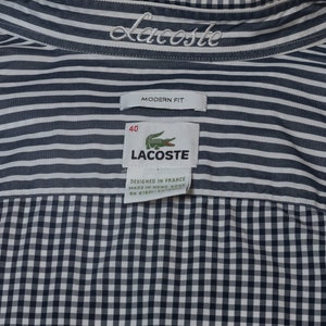 May include: Close-up of a Lacoste shirt with a striped upper section and a checkered lower section. The shirt features the Lacoste logo and brand name. The tag reads "Modern Fit" and "Designed in France".