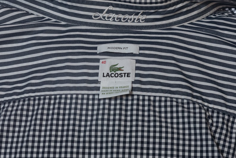 May include: Close-up of a Lacoste shirt with a striped upper section and a checkered lower section. The shirt features the Lacoste logo and brand name. The tag reads "Modern Fit" and "Designed in France".