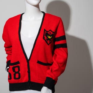 May include: Red knitted cardigan with black trim and a black and yellow embroidered logo on the front. The cardigan has two black pockets and a black number "58" on the left side.