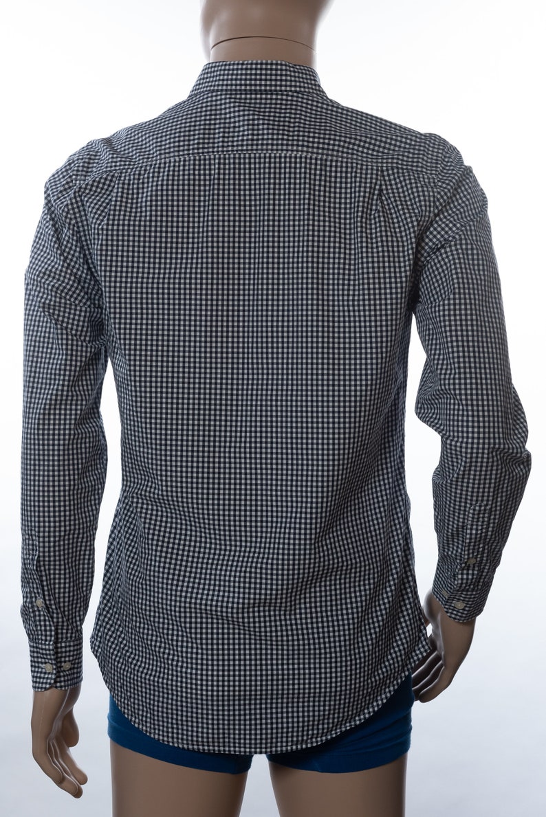 May include: A long-sleeved button-down shirt in a navy and white gingham pattern. The shirt is displayed on a mannequin, showcasing the back view. The cuffs have button closures. The shirt is made of a lightweight fabric.