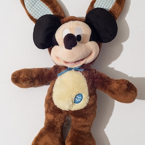 May include: Brown plush Mickey Mouse toy wearing a brown bunny costume with blue and white checkered ears. The toy has a yellow patch on its chest with a blue Aquarius symbol.
