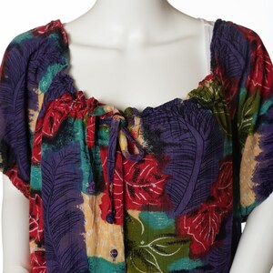 May include: A colourful, loose-fitting blouse with a tie at the neckline and a button-up front. The fabric features a vibrant abstract floral print in shades of red, purple, green, and yellow.