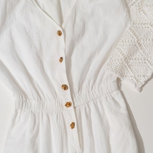 Può includere: Abito bianco in cotone con un'applicazione di pizzo all'uncinetto sulle maniche. L'abito ha una scollatura a V e una chiusura con bottoni sul davanti. L'abito ha una vita stretta e una gonna ampia.