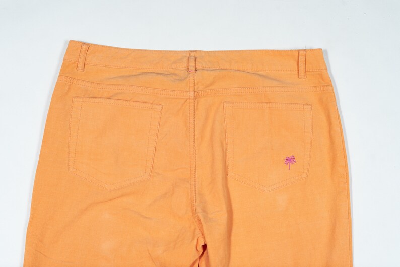 May include: A pair of orange cotton pants with two back pockets and a small pink palm tree embroidered on the right back pocket.