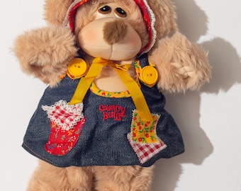 May include: A plush brown teddy bear wearing a red and white gingham bonnet and a blue denim dress with patchwork accents. The dress has yellow straps and two yellow buttons. The bear's dress has the text "Country Bear" embroidered on the front.