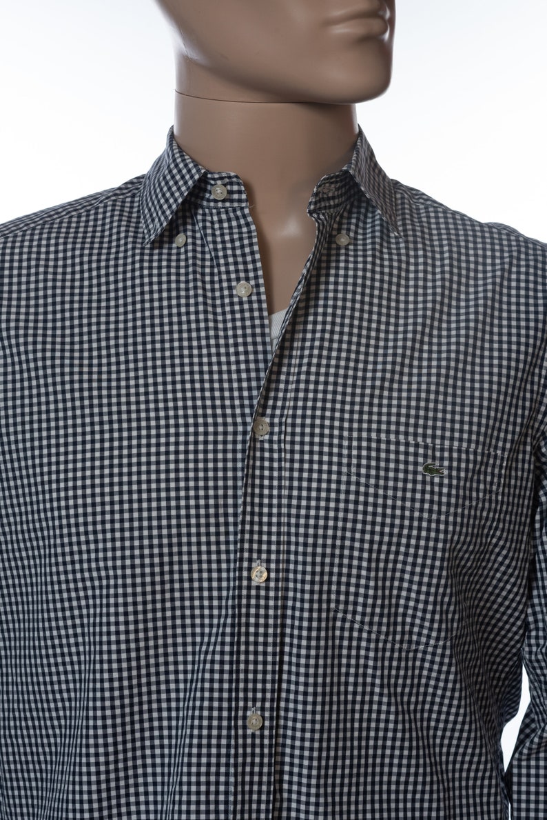 May include: A button-down shirt with a navy and white gingham pattern. The shirt has a chest pocket and a small green logo. The collar is buttoned down. The shirt is on a mannequin.