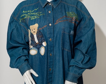 Può includere: Una camicia di jeans blu con maniche lunghe e chiusura con bottoni. La camicia presenta un ricamo di un orsacchiotto che indossa una giacca di jeans e jeans, circondato da alberi.