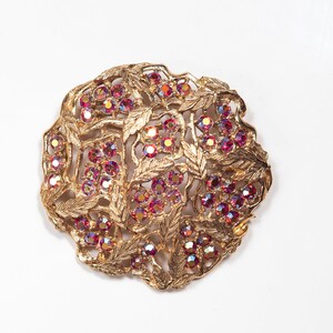 Può includere: Un broche dorato con un delicato design di foglie e strass rossi iridescenti.
