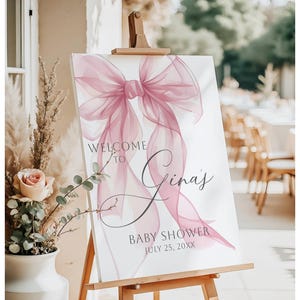 May include: A welcome sign for a baby shower, featuring a large, translucent pink bow design. The sign reads "Welcome to Gina's Baby Shower July 25, 20XX" in elegant script and sans-serif fonts. The sign is displayed on a wooden easel.