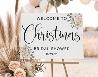 May include: A white sign on a wooden easel with the words "Welcome to Christinas Bridal Shower 8.29.21" in black script. Floral arrangements in peach and green adorn the corners. The sign is set up outdoors.