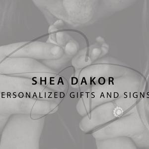 May include: Monochrome image featuring the text "SHEA DAKOR PERSONALIZED GIFTS AND SIGNS." The image shows hands holding a baby's foot and a ring with a floral design.