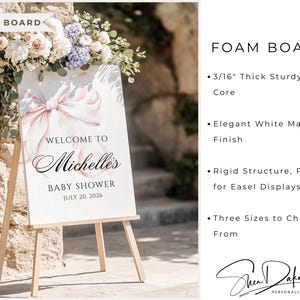 May include: A white foam board sign on a wooden easel, with a pink bow and the text "Welcome to Michelle's Baby Shower July 20, 2026." The sign is surrounded by flowers.