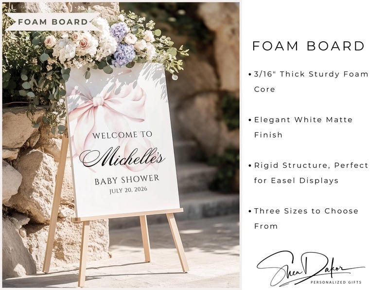 May include: A white foam board sign on a wooden easel, with a pink bow and the text "Welcome to Michelle's Baby Shower July 20, 2026." The sign is surrounded by flowers.