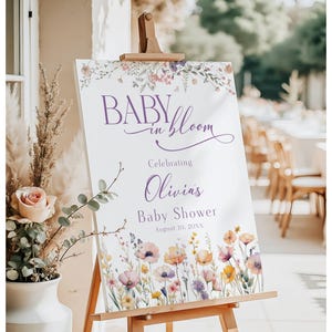May include: A white sign on a wooden easel with the text "BABY in bloom" in purple script, celebrating Olivia's baby shower. The sign is decorated with watercolor floral illustrations in pink, orange, and purple.