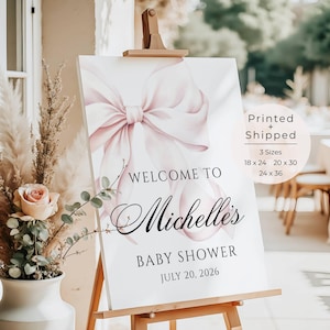 May include: A baby shower welcome sign on an easel. The sign features a large pink bow illustration and the words "Welcome to Michelle's Baby Shower July 20, 2026". The sign is available in 3 sizes: 18 x 24, 20 x 30, and 24 x 36 inches.