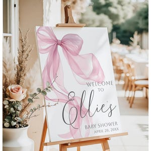 May include: A white sign on a wooden easel with a watercolor-style pink bow and the words "Welcome to Ellie's Baby Shower" in elegant script. The date is April 20, XXXX. The sign is displayed outdoors.