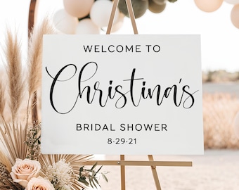 May include: A white sign on a wooden easel with the text "WELCOME TO Christina's BRIDAL SHOWER 8.29.21" in black script. The sign is decorated with flowers and pampas grass, with balloons in the background.