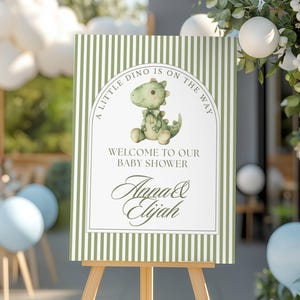 May include: A baby shower welcome sign on a wooden easel. The sign has a green and white striped background with a cartoon dinosaur illustration. Text reads "A Little Dino Is On The Way" and "Welcome To Our Baby Shower" with the names "Anna & Elijah".