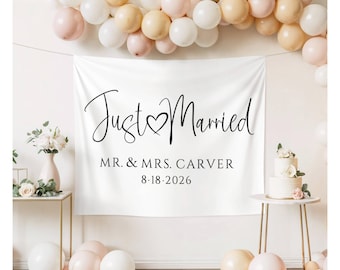 May include: A white banner with the words "Just Married" in elegant black script, with a heart symbol. Below, it reads "MR. & MRS. CARVER 8.18.2026". The banner is decorated with balloons and floral arrangements, suggesting a wedding theme.
