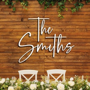 May include: A white wall-mounted sign featuring the text The Smiths in a cursive script font. The sign is displayed against a horizontal brown wooden plank background, positioned above two white chairs and a floral arrangement of white flowers.