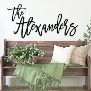 May include: A black wall-mounted sign featuring the words "the Alexanders" in a cursive script font. The sign is displayed above a wooden bench with a green throw blanket, a white pillow, a textured beige pillow, and a basket of flowers.