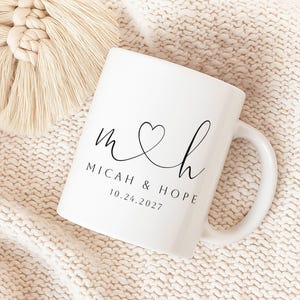 May include: A white ceramic mug featuring black printed text that reads M and H with a heart symbol connecting the letters, followed by the names MICAH & HOPE and the date 10.24.2027 in a clean, minimalist sans-serif and script font style.