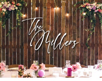 May include: A white script sign reading The Millers mounted on a dark brown vertical wooden plank wall. The display is decorated with hanging floral arrangements of pink, white, and purple flowers with green vines and string lights.