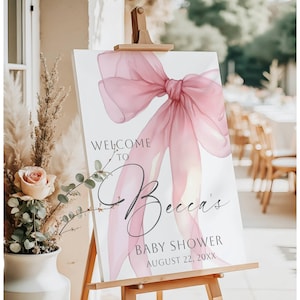 May include: A white rectangular welcome sign for a baby shower featuring a large, translucent, dusty pink ribbon bow graphic. The text reads: Welcome to Becca's Baby Shower August 22, 20XX. The sign is displayed on a wooden easel stand.