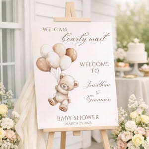 May include: A white baby shower welcome sign on an easel. The sign features a watercolor illustration of a teddy bear holding balloons. Text includes "We can bearly wait", "Welcome to Jonathan & Gianna's", and "Baby Shower March 25, 2026".