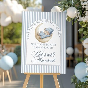 May include: A baby shower welcome sign on a wooden easel. The sign has a blue and white striped background with a graphic of a teddy bear sleeping on a crescent moon. Text reads "Our Little Cuddle Bug Is On His Way" and "Welcome to Our Baby Shower" with the names "Victoria & Maverick".