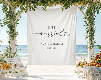 May include: A white banner with the words "Just Married" in elegant script, along with the names Jackie & Daniel and the date 11-15-2026. The banner is part of a beach wedding setup, with floral arrangements and a view of the ocean.