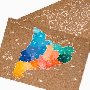May include: Two corkboard maps of Catalonia, Spain, with colorful regions and white outline details. The maps feature place names like Barcelona and Girona. The corkboard has a natural brown color, and the maps are layered.