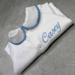 Puede incluir: Mono blanco para bebé con ribete azul alrededor del cuello y el nombre "Casey" bordado en azul en la parte delantera.