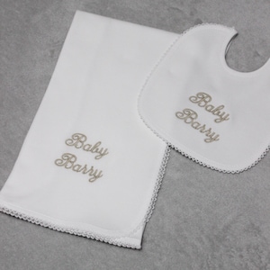 May include: A white baby blanket and bib set with a gold embroidered name "Baby Barry" on both items. The blanket and bib have a white lace trim.